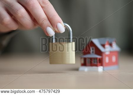 Hand Of Woman Holding Iron Door Lock Against Blurred Small Model Of Cottage House. Two-story Buildin