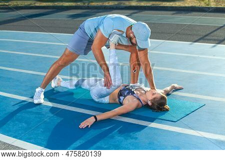 Instructor And Athlete Runner On The Track. The Trainer Stretches The Gluteal Muscles Of The Girl. F