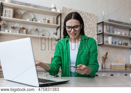 Worried Young Woman Sitting At Home In Kitchen In Front Of Laptop And Looking Shocked At Credit Card