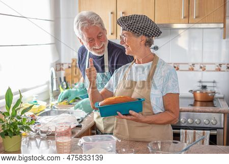 Happy Senior Couple In The Home Kitchen While Wife Holds A Freshly Baked Homemade Plumcake And Husba