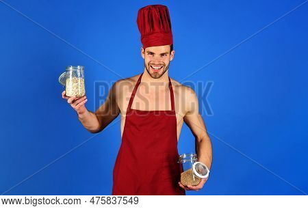 Smiling Male Chef In Burgundy Hat And Apron Holds Glass Jars With Groats And Cereals. Professional C