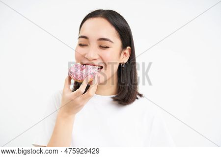 Close Up Of Smiling Pleased Asian Woman, Eats Glazed Pink Doughnut, Enjoys Eating Tasty Donnut, Whit