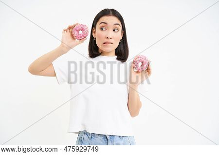 Image Of Asian Girl Holds Two Doughnuts, Thinking Of Breaking Her Diet, Looks With Doubt At Donnuts,