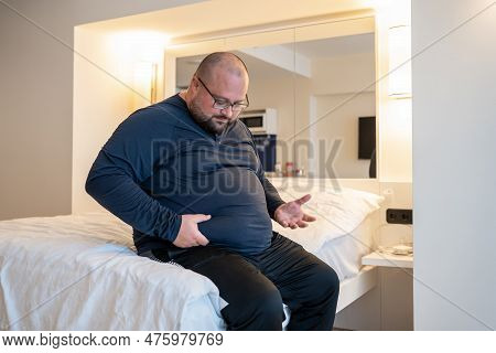 Huge Man Suffering From Extra Weight Touching Stomach Sitting On Bed In Bedroom. Overweight Bearded 