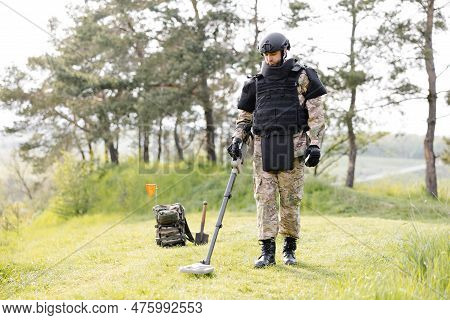 A Man In A Military Uniform And Bulletproof Vest Works In The Forest With A Metal Detector. A Minesw