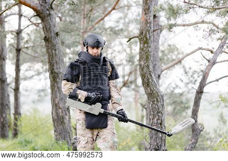 A Man In A Military Uniform And Bulletproof Vest Works In The Forest With A Metal Detector. A Minesw