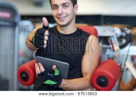 sport, bodybuilding, technology, gesture and people concept - happy young man with tablet pc computer showing thumbs up in gym