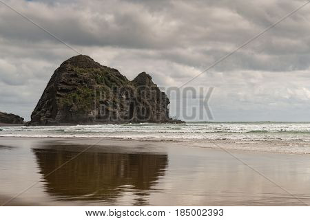 Auckland New Zealand - March 2 2017: Closeup of Rabbit rock is mirrored on wet sandy Piha Beach under stormy sky and surrounded by Tasman Sea surf.