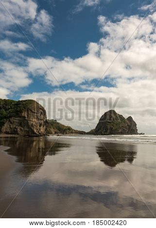 Auckland New Zealand - March 2 2017: Closeup of Rabbit rock is mirrored on wet sandy Piha Beach under blue cloudy sky and surrounded by Tasman Sea surf. Forested cliff.