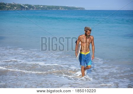 Attractive bearded curly man with a surfboard is walking on a beach located on the right side of the frame