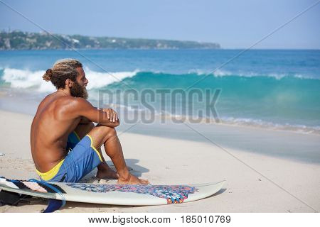 Attractive bearded curly man with a surfboard is seating with his back on a sand and looking to the ocean located on the left side of the frame
