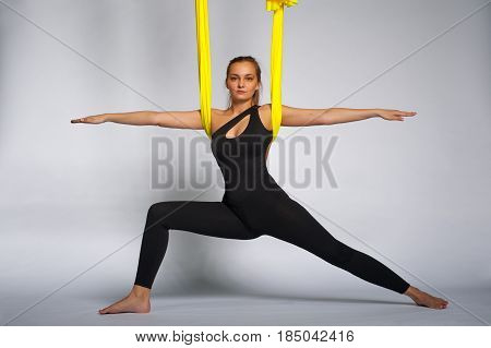 Young beautiful girl practicing aerial yoga in studio. Attractive blonde woman doing warrior yoga pose with yellow hammock. Body balance and antigravity training. Wellness and body care concept.