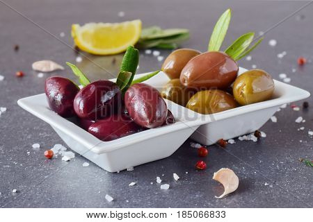 Black and green olives in a white plate on a grey abstract background with lemon, garlic, spices. Mediterranean food. Healthy eating concept