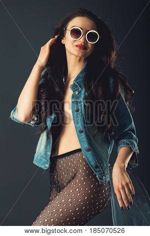 gorgeous girl in a fashionable white sunglasses, dressed in a blue jeance jacket and transparent tights without panties. posing standing near stylish transparent armchair. side view, waist portrait