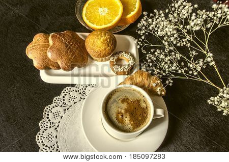 Koffee in white cup dishes with confectionery cakesa glass saucer with orange slices on black wooden table
