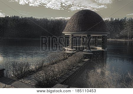 A sepia photograph of a gazebo in the woods on a lake during winter time.