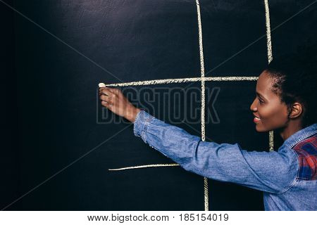 Afroamerican young smiling girl play tic tac toe game with white chalk on black board, close up. Fun, entertainment, leisure, strategy concept.