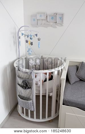newborn lies in the round white bed with mobile and toys