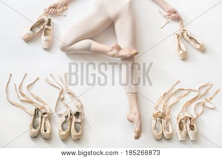 Pretty ballerina lies in passe on the white floor in the studio. She wears a light dance wear and holds ribbons of the shoes in her hands. On the sides of her legs there are ballet shoes. Horizontal.