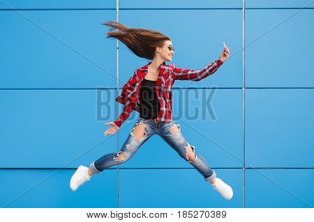 Fashion portrait of pretty smiling and jumping woman in sunglasses with smartphone against the colorful blue wall. Copyspace.
