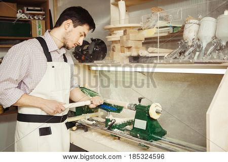 Young turner engages in wood carving on lathe for wood. Wooden blank clamped in metal centers of machine-tool. Worker in overalls holding hand milling cutters.