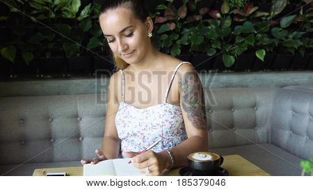 Young prety woman with tattoo writing in her journal, diary. Caucasian model with long hair. Cup of coffee and smartphone on the table,