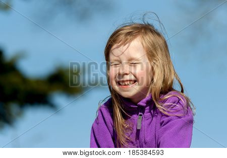 portrait of a little girl in a lilac jacket on a blue sky background on a sunny day, the child laughs and closed his eyes, a wide smile