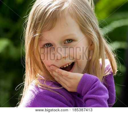 a portrait of a little girl in a lilac jacket on the background of green leaves in the garden, her hand is covered with a mouth half, a little gossip, a secret to tell,  long blond wheat hair,