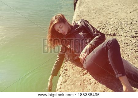 beautiful woman in jeans and jacket lies on the breackwater. And touch the water