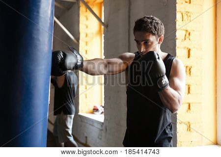 Photo of young strong boxer training in a gym. Looking aside. Standing near punchbag.