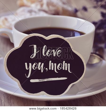 closeup of a black cloud-shaped signboard with the text I love you mom written in it, next to a cup of coffee on a table set for breakfast with some viennoiseries in a plate in the background