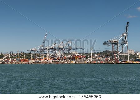 Auckland New Zealand - March 3 2017: The container terminal with its cranes and boxes under blue sky and behind greenish ocean water. War Memorial Museum on hill in background.