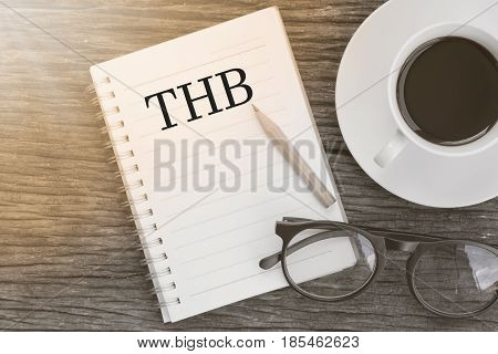 Concept THB message on notebook with glasses pencil and coffee cup on wooden table.