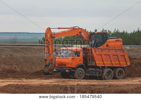 Heavy excavator loading dumper truck on road construction among fields, telephoto