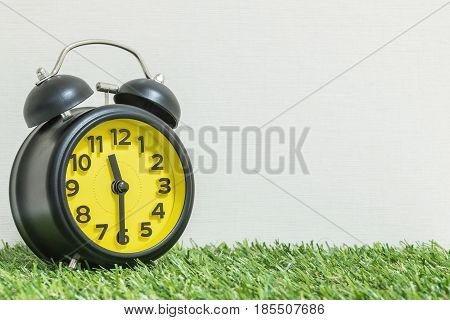 Closeup black and yellow alarm clock for decorate show half past eleven o'clock or 11:30 a.m. on green artificial grass floor and cream wallpaper textured background with copy space