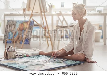 Beautiful and young woman sitting on the floor in a bright workshop and paints with paint on canvas.