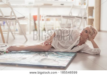 Beautiful young artist, is on the floor in a bright Studio and posing next to the canvas