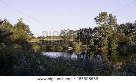 Landscape scene of a small river shoreline with trees foliage and reflections in the water near Windsor, Ontario with the moon visible on a sunny late afternoon in September.