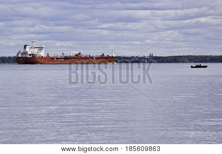 Notre Dame de Ile Perrot, Quebec - June 20, 2014 -- Wide landscape view of a very large red cargo ship and a very small boat motor boat in Lake St-Louis in Notre Dame de Ile Perrot, Quebec with trees foliage in the background on a sunny day in June.