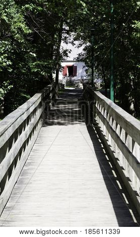 Vertical perspective view of a walking bridge leading to a white house with red shutter window at the end, shot in Notre Dame de Ile Perrot, Quebec on a sunny day in June.