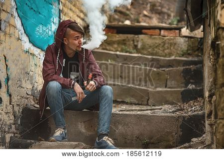 Vape man. Photo of a handsome young white guy in the hood vaping and letting off steam from an electronic cigarette on an old broken concrete staircase. Lifestyle.