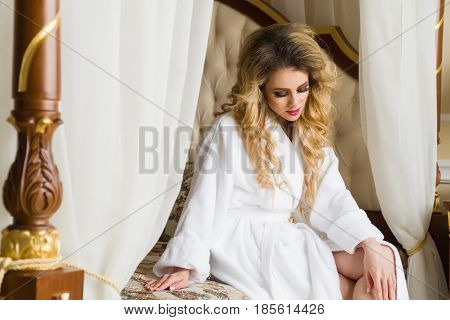 Beautiful seductive woman flirting with the camera sitting on a bed in White bathrobe looking up with a coquettish look.