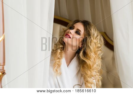 Beautiful seductive woman flirting with the camera sitting on a bed in White bathrobe looking up with a coquettish look.