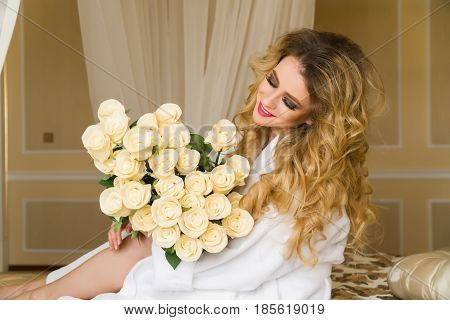 Beautiful seductive woman flirting with the camera is sitting on the bed with a big bouquet of white roses in White bathrobe looking up with a coquettish look.