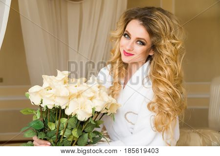 Beautiful seductive woman flirting with the camera is sitting on the bed with a big bouquet of white roses in White bathrobe looking up with a coquettish look.