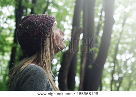 horizontal side view portrait of Caucasian young woman with long blonde hair and colorful wool hat standing in a green forest and looking up