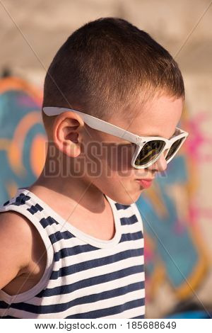 funny small kid wearing sunglasses and sailor shirt on graffiti background in summer evening