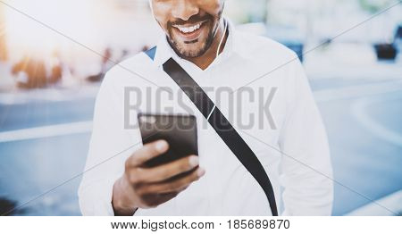 Happy american African man using smartphone outdoor.Portrait of young black cheerful man texting a sms message while listining to music.Blurred background, horizontal cropped.Flare effects