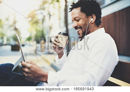 Smiling African American man in earphones making video call via electronic touch pad with take away coffee in hand.Concept of guy using Internet-enabled electronic device outdoor.Blurred background