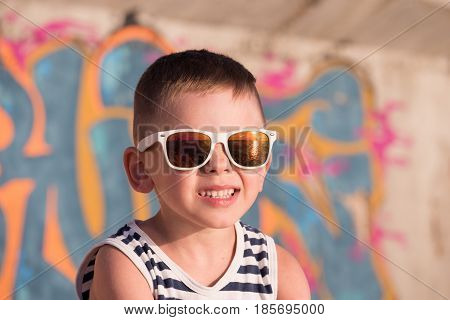 funny small kid wearing sunglasses with sea sunset reflection in it and sailor shirt on graffiti background lit by warm summer light